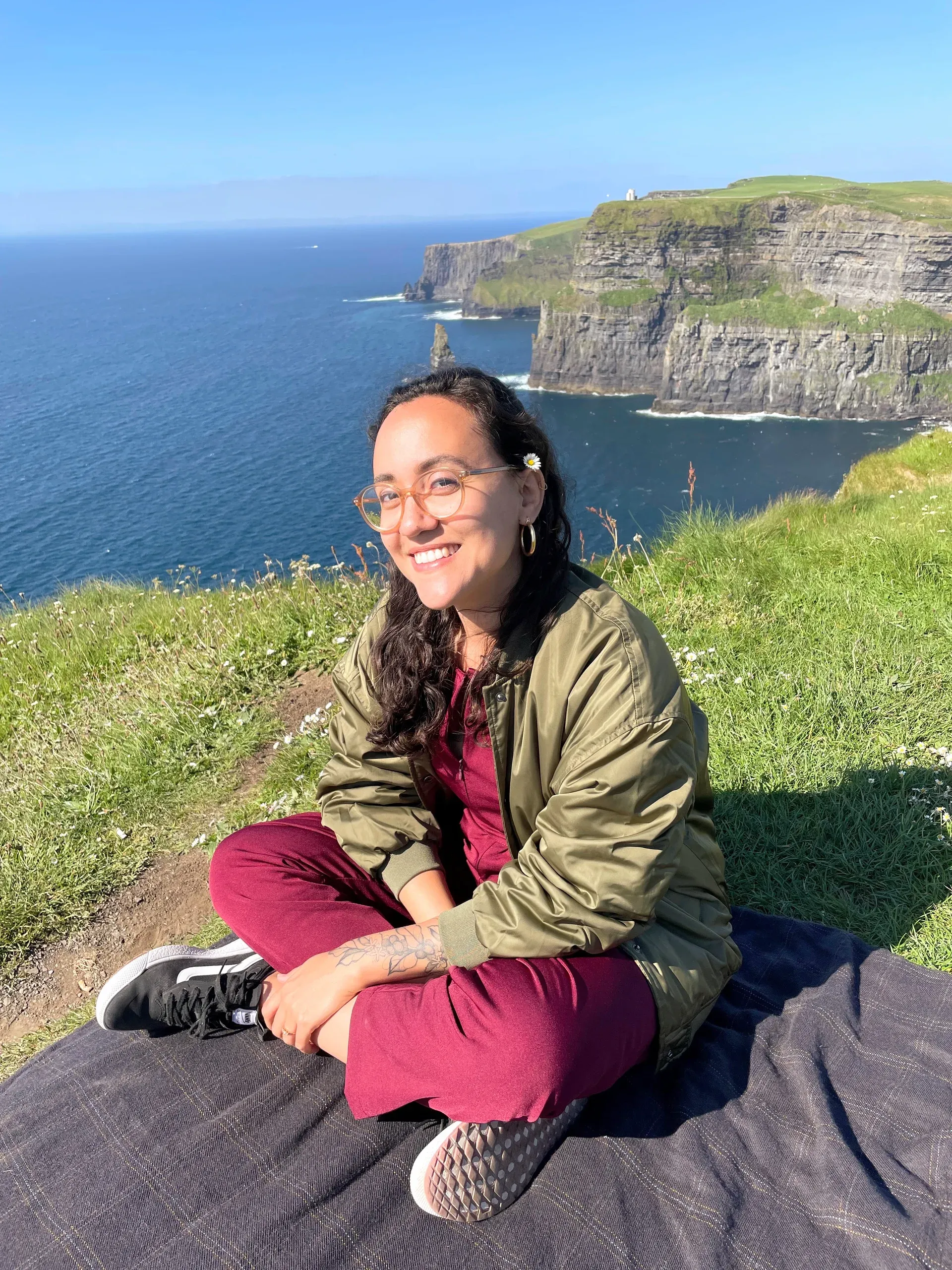Renata smiles and sits on the grass at the Cliffs of Moher - Ireland.