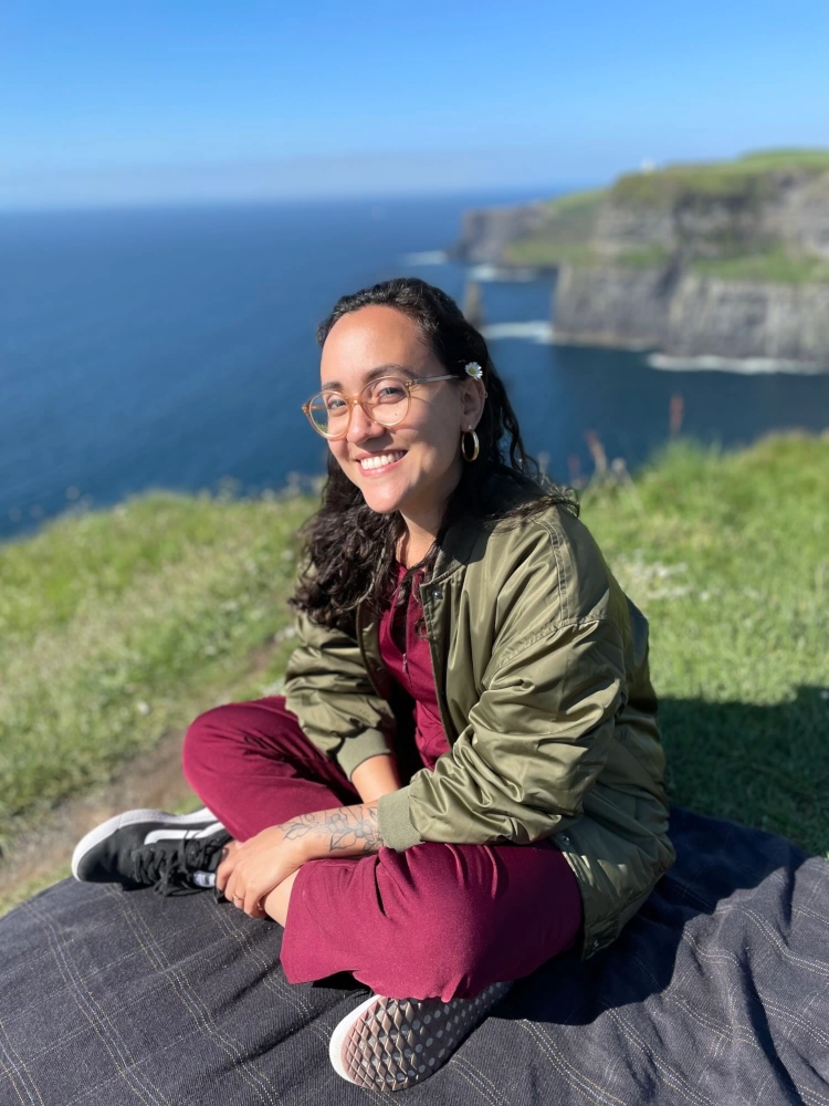 Renata smiles and sits on the grass at the Cliffs of Moher - Ireland.